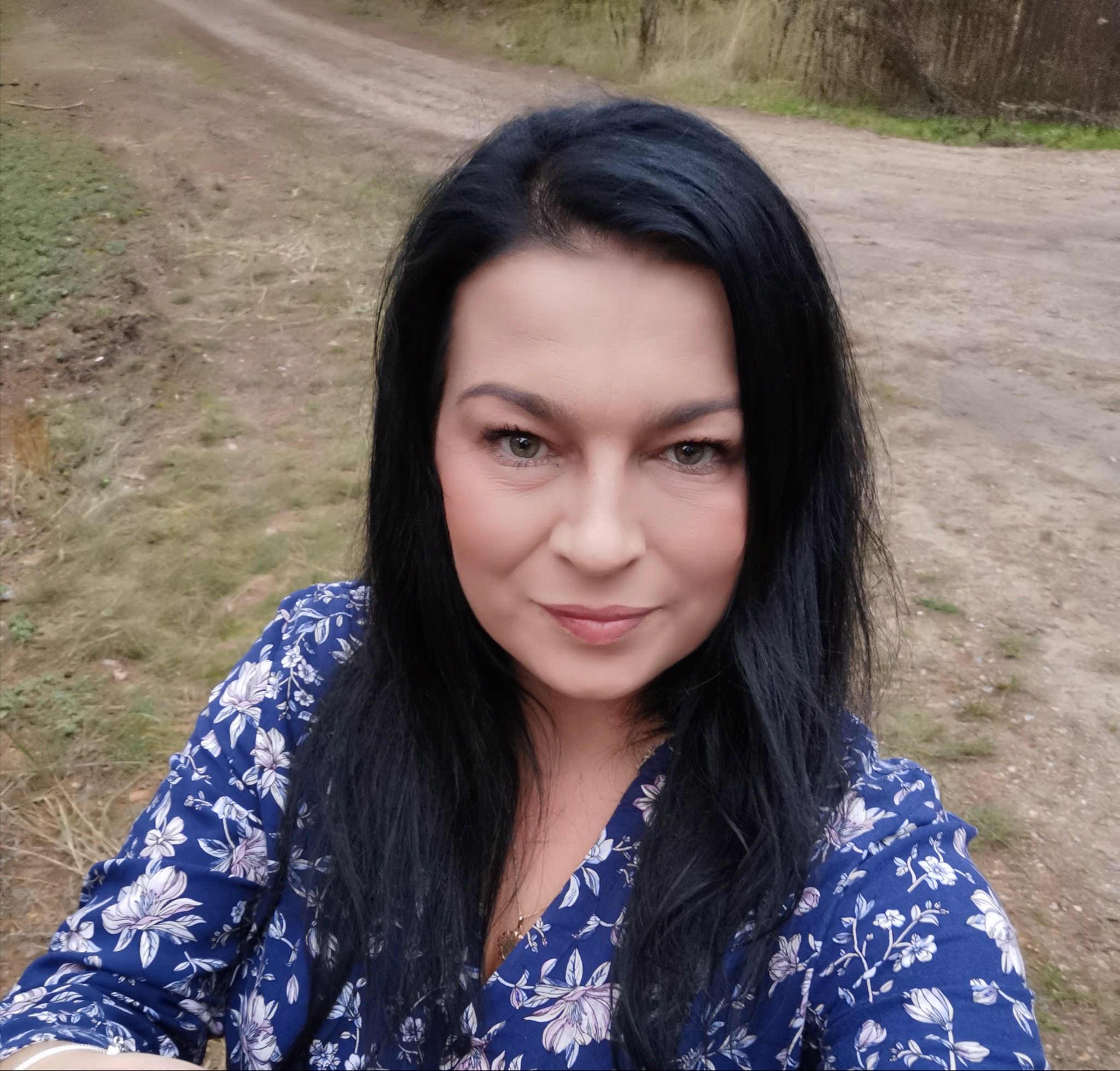 Portret kobiety z czarnymi włosami i niebieskiej sukience w kwiaty na tle polnej drogi.