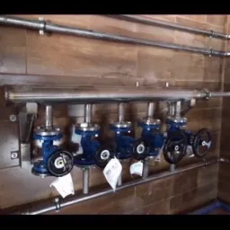 Rząd czterech niebieskich zaworów z czarnymi pokrętłami, podłączonych do rury ze stali nierdzewnej, zamocowanych na ścianie z płytek imitujących drewno, z widocznymi etykietami na zaworach.