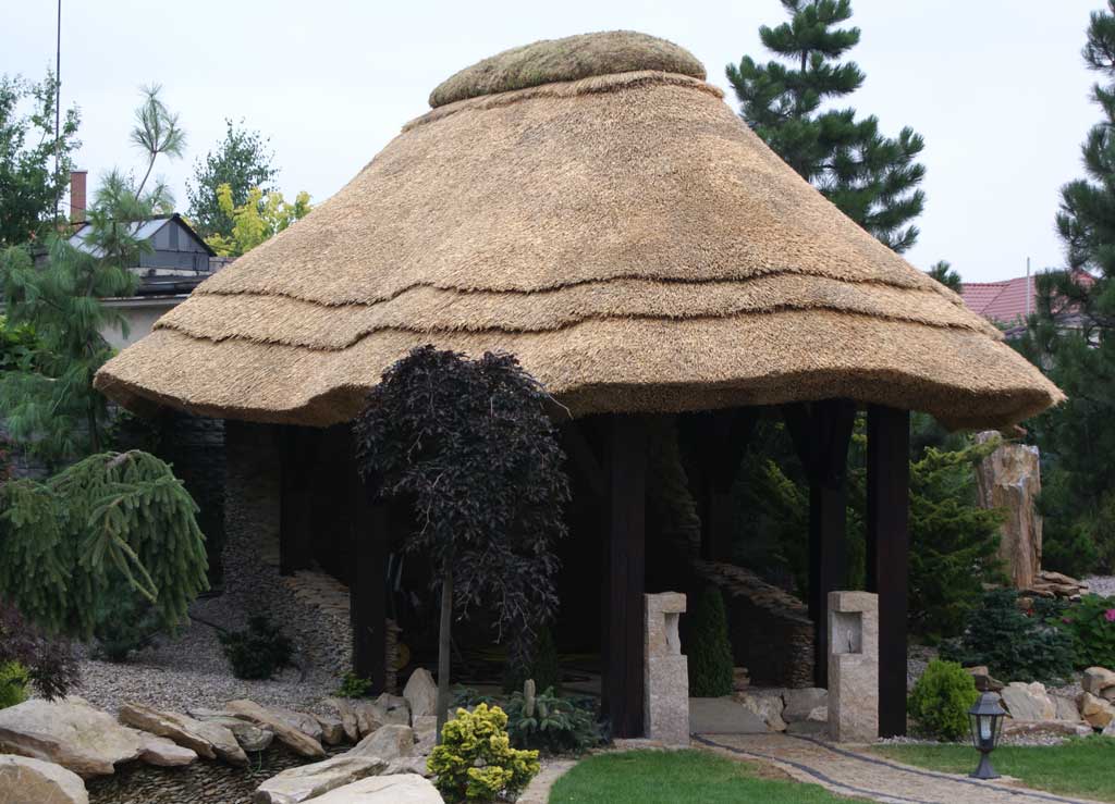 Altana kryta strzechą z widoczną strukturą ułożenia słomy, otoczona kamieniami i zielenią w ogrodzie.