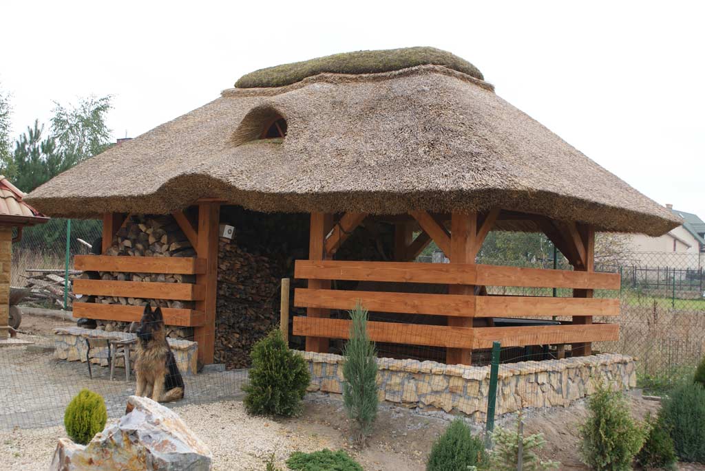 Altana z dachem krytym strzechą, drewniane słupy i balustrady, kamienna podmurówka, zapas drewna wewnątrz, pies owczarek niemiecki siedzący z przodu.