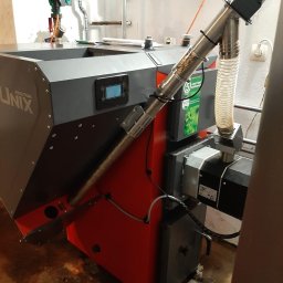 Mateusz Strzelecki - VIP Energy - Kocioł na pellet Kolton Unix z podajnikiem, widoczne rury doprowadzające paliwo i panel sterowania, tło instalacji hydraulicznej.