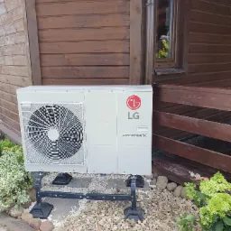 Biała jednostka zewnętrzna z logo LG Therma V, zamontowana na czarnym metalowym stelażu, ustawiona na żwirowym podłożu przy drewnianym budynku z oknem i schodami.