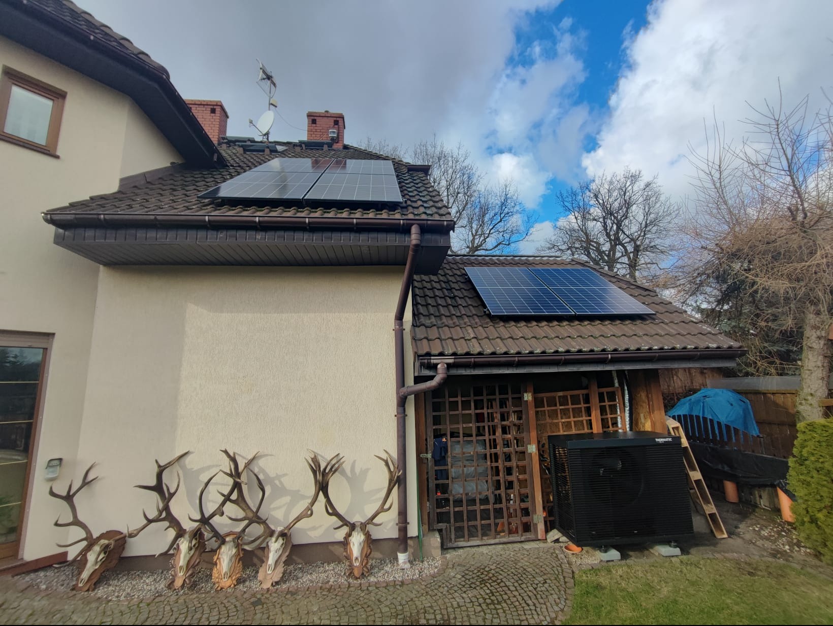 Dom z panelami słonecznymi na dachu i głowami jelenia pod ścianą, obok czarna jednostka zewnętrzna pompy ciepła.