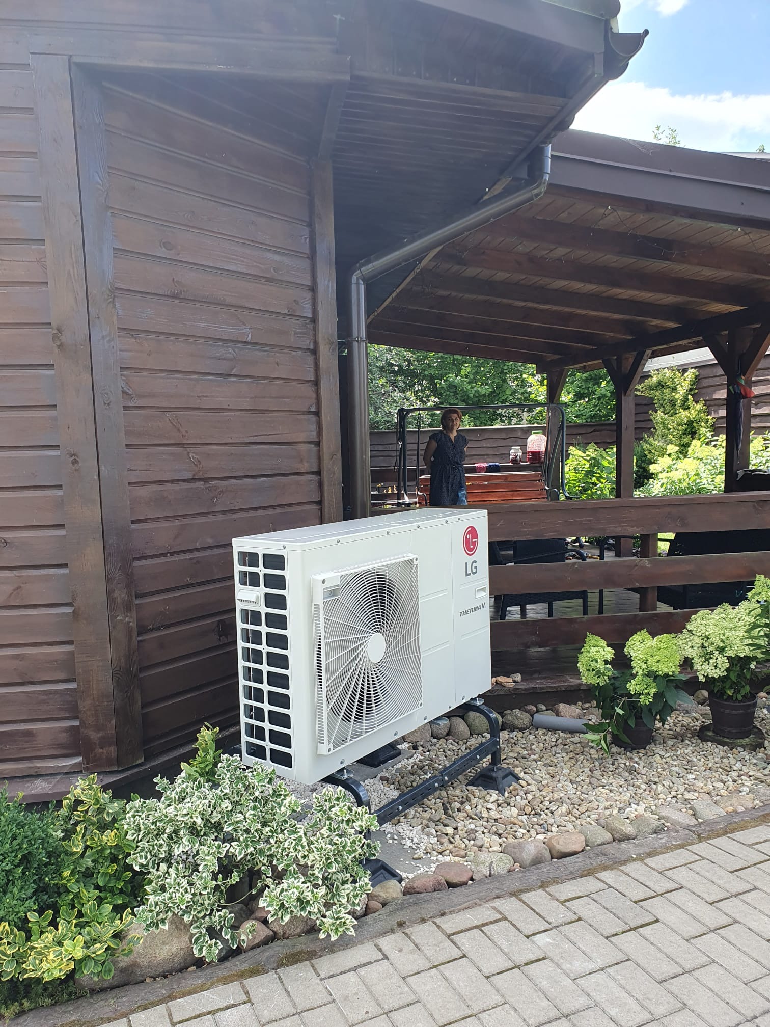 Biała jednostka zewnętrzna z logo LG Therma V, zamontowana na metalowym stelażu przy drewnianej altanie, otoczona ozdobnymi krzewami i kamieniami.