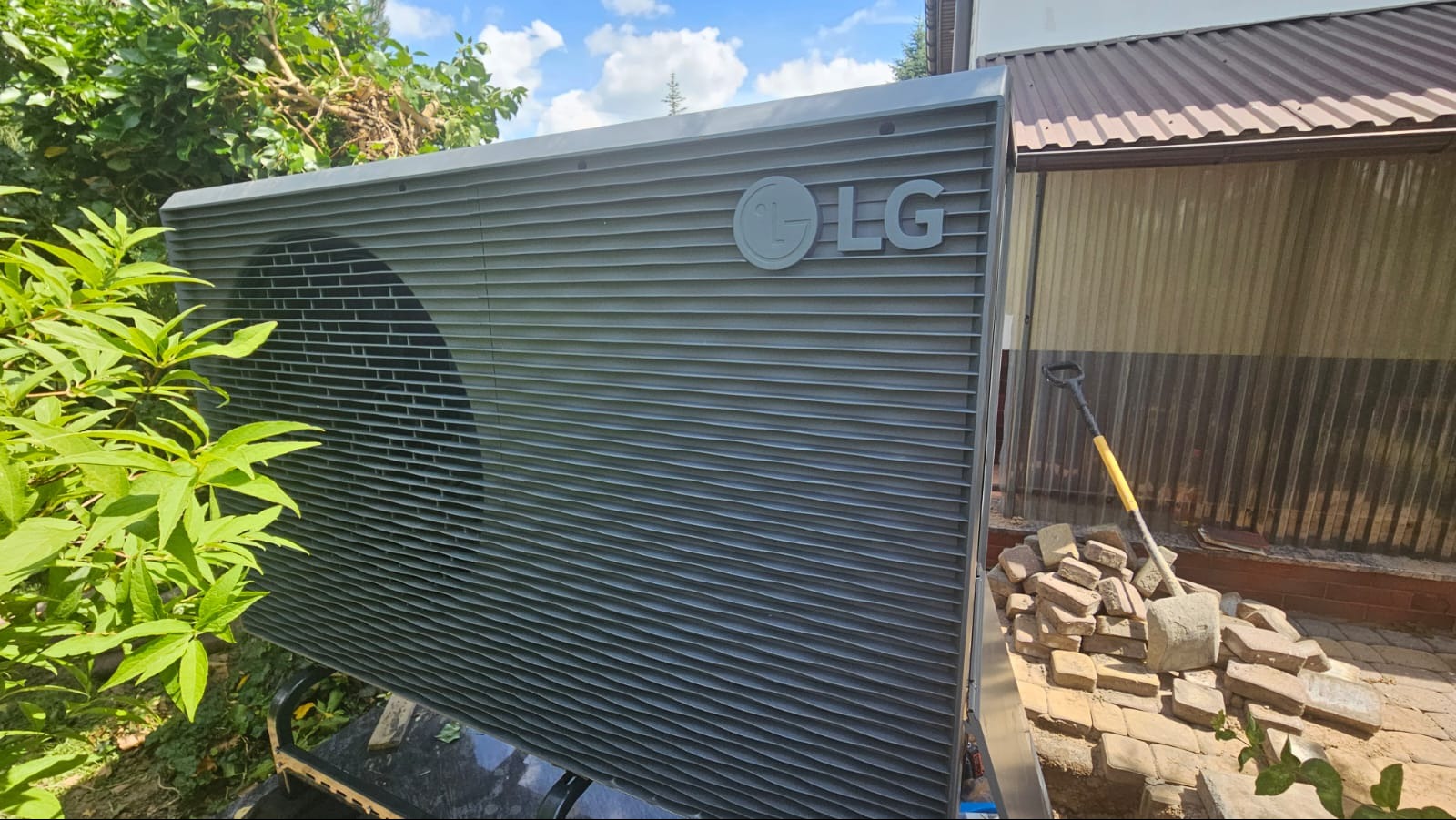 Szara jednostka zewnętrzna z logo LG, częściowo zasłonięta zielonymi liśćmi, z widoczną łopatą i stertą cegieł w tle.