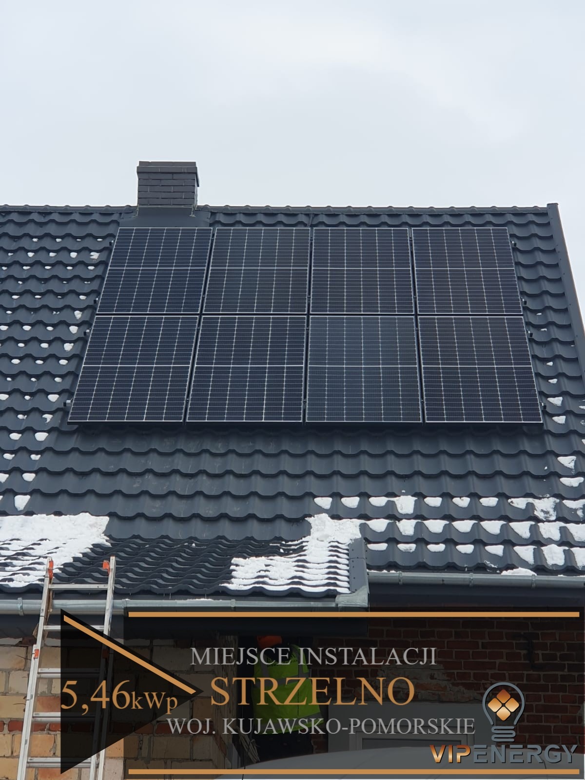 Panel słoneczny o mocy 5,46 kWp zainstalowany na ciemnoszarym dachu pokrytym śniegiem, widoczna drabina oparta o ceglany budynek, miejscowość Strzelno, województwo kujawsko-pomorskie, logo firmy...