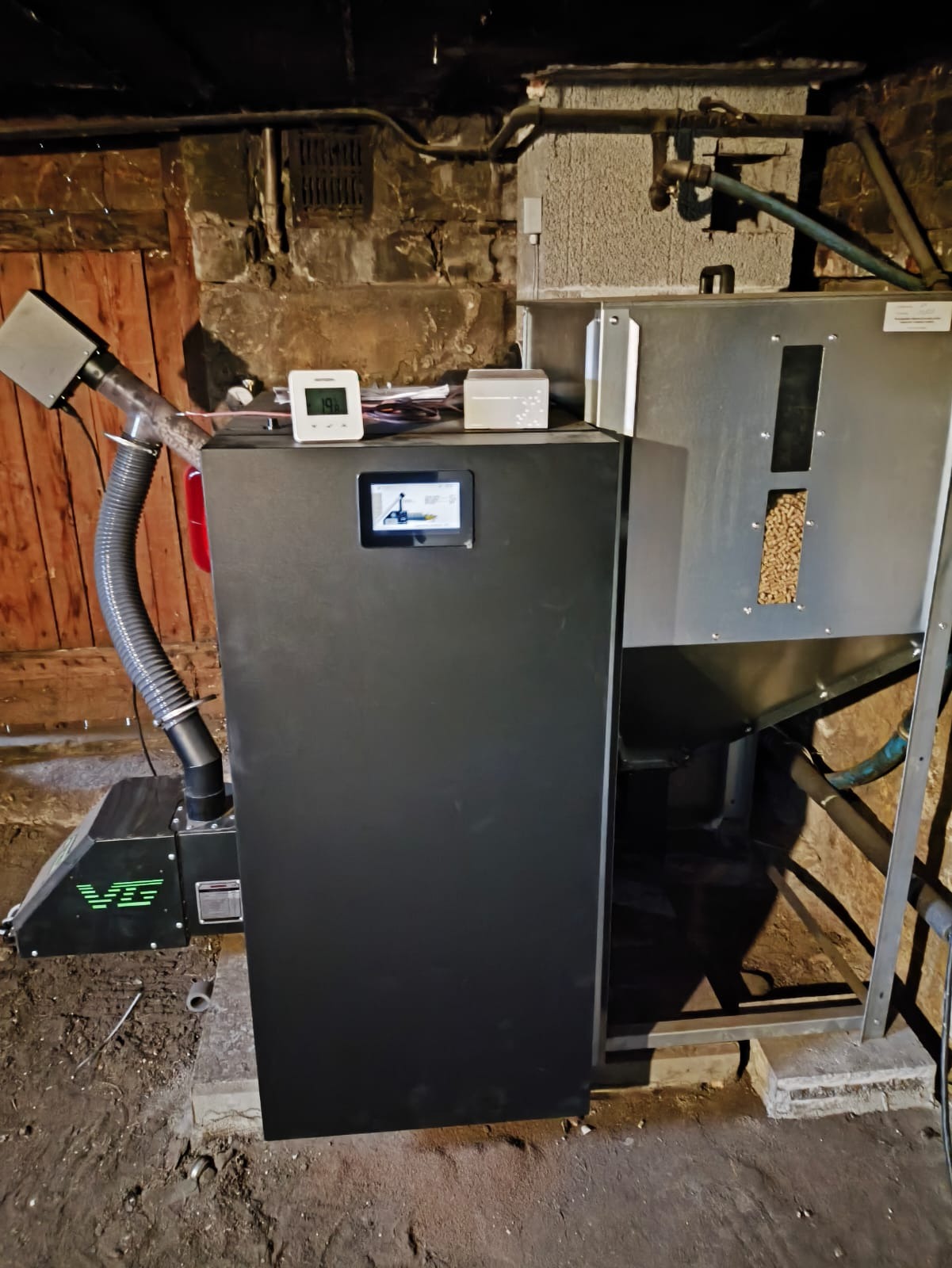 Nowoczesny piec na pellet w kotłowni ze ścianami z kamienia i cegły, z widocznym zasobnikiem na pellet i panelem sterowania.