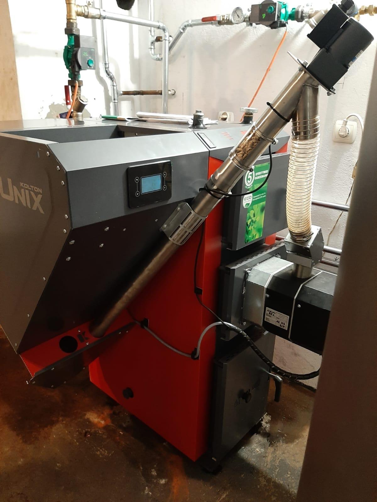 Kocioł na pellet Kolton Unix z podajnikiem, widoczne rury doprowadzające paliwo i panel sterowania, tło instalacji hydraulicznej.