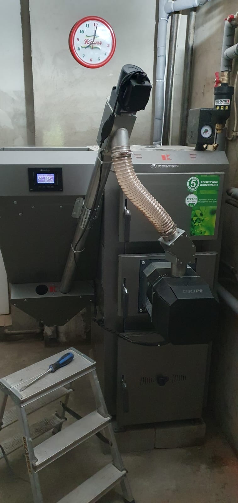 Szary kocioł grzewczy marki Kolton z podajnikiem, widoczny na ceglanych podstawach w pomieszczeniu, z zegarem ściennym w tle i śrubokrętem na aluminiowej drabinie.