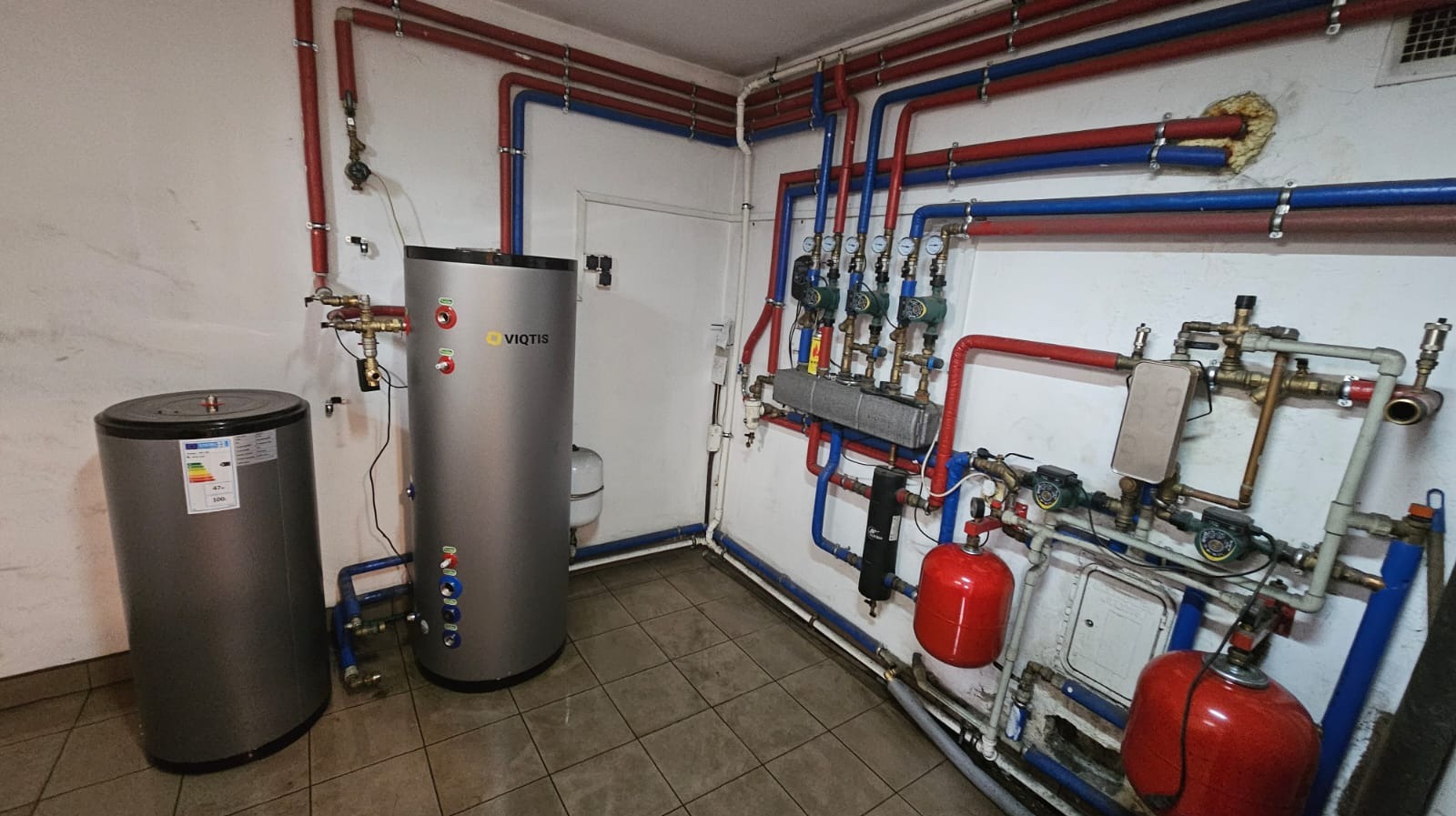 Kotłownia z dwoma zbiornikami akumulacyjnymi, licznymi rurami w kolorach czerwonym i niebieskim oraz osprzętem hydraulicznym na ścianie, widoczne logo Viqtis na jednym ze zbiorników.