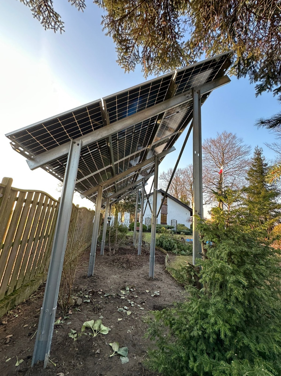 Instalacja fotowoltaiczna naziemna z metalową konstrukcją wsporczą w ogrodzie, widok z dołu, w tle dom i drzewa.