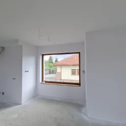 Wnętrze pokoju po świeżym pomalowaniu na biało, z widocznymi przewodami elektrycznymi wystającymi z sufitu i oknem z widokiem na dom z czerwonym dachem.