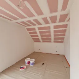 Poddasze w trakcie prac wykończeniowych: widoczne płyty kartonowo-gipsowe na skośnym suficie, spoiny wypełnione masą, zabezpieczona folią podłoga i stojące obok ściany wiaderka z farbą.