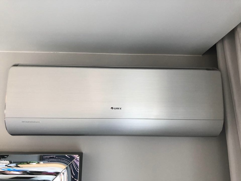 Srebrny klimatyzator marki Gree zamontowany na szarej ścianie w pomieszczeniu z zasłoną i widocznym fragmentem telewizora.