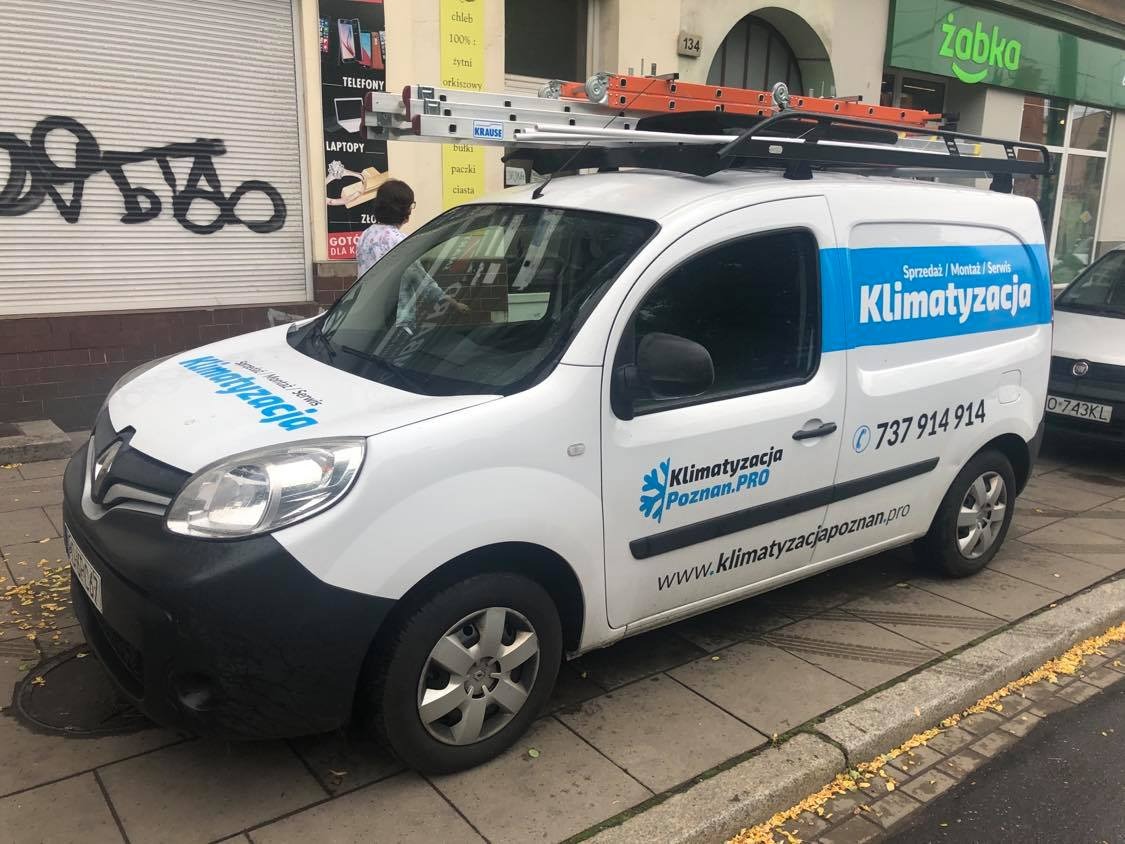 Biały samochód dostawczy z logo firmy Klimatyzacja Poznań.PRO i numerem telefonu, zaparkowany na chodniku przed sklepem Żabka, z drabiną na dachu.