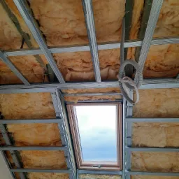 Poddasze z oknem dachowym w trakcie izolacji wełną mineralną, widoczne metalowe profile konstrukcyjne i przewód elektryczny w peszlu.
