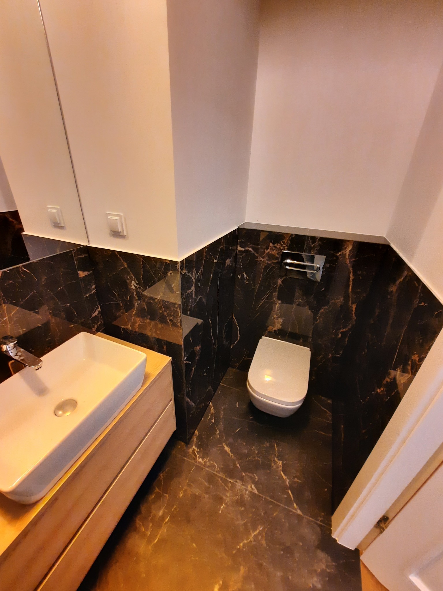 Nowoczesna łazienka z umywalką nablatową na drewnianej szafce i podwieszaną toaletą, wykończona czarnym marmurem z wyraźnym żyłowaniem.
