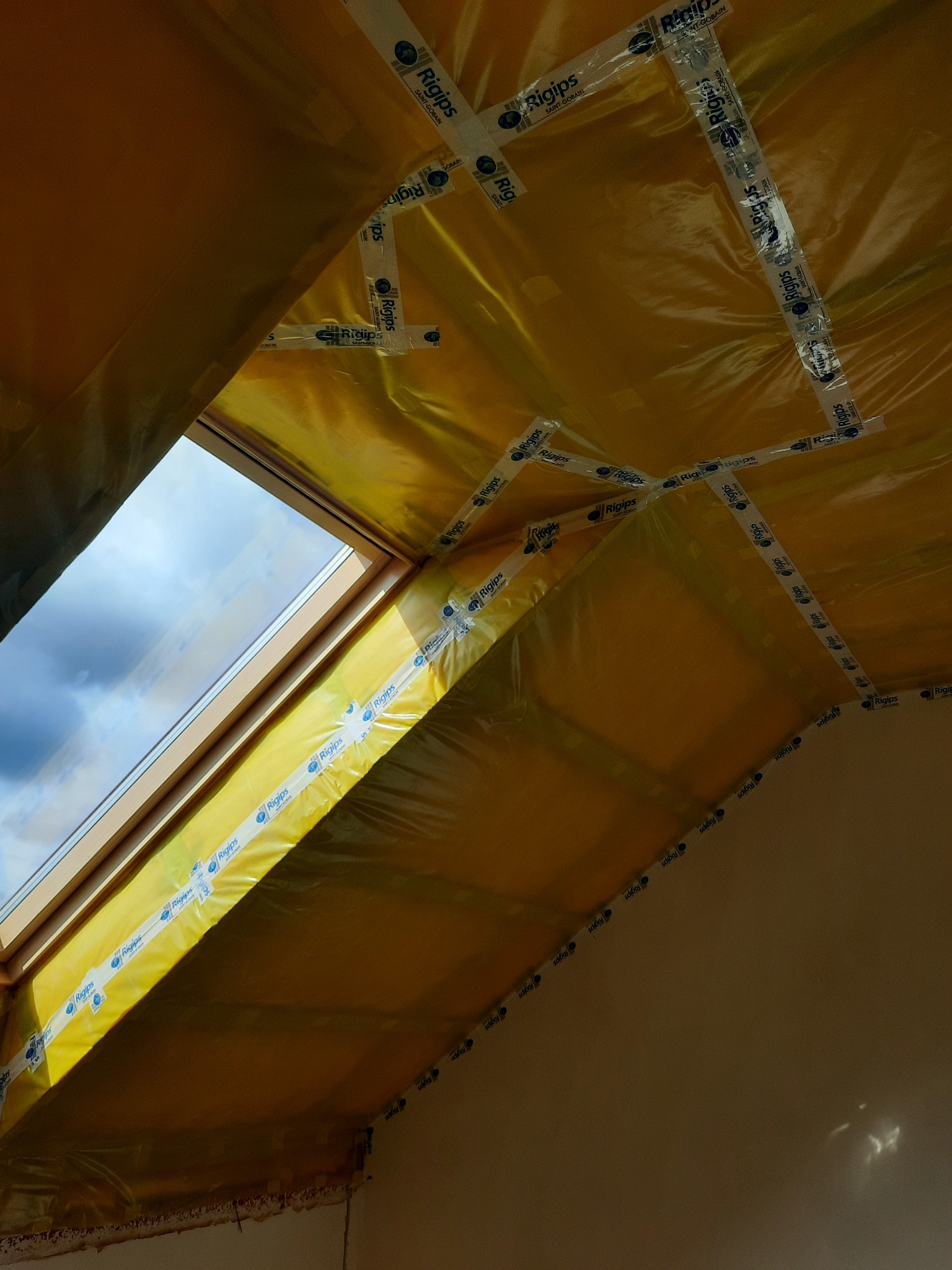Poddasze z oknem dachowym w trakcie izolacji, widoczna żółta folia paroizolacyjna przymocowana taśmą marki Rigips, fragment białej ściany i błękitne niebo za oknem.