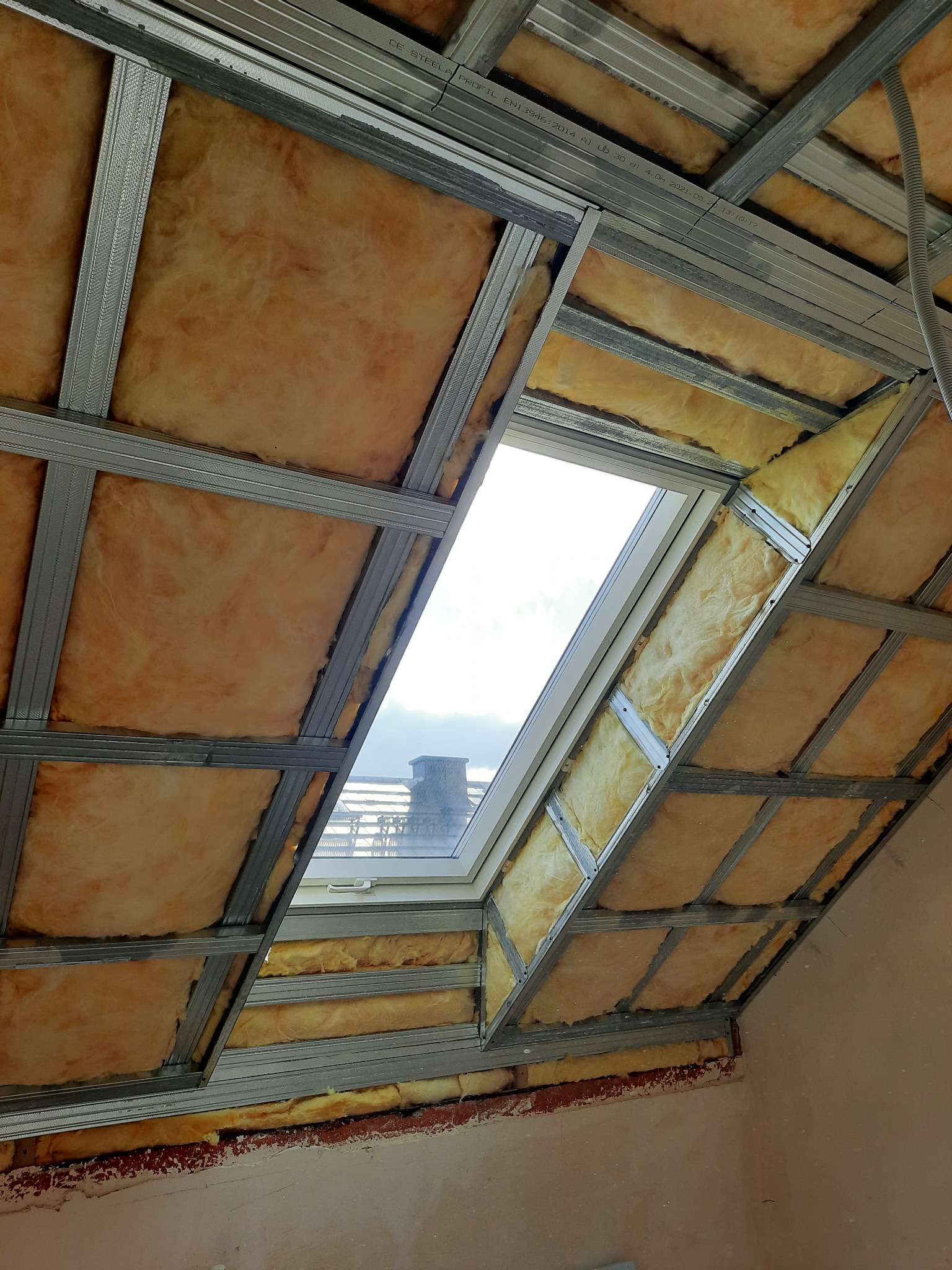 Montaż izolacji z wełny mineralnej na poddaszu, widoczne metalowe profile konstrukcyjne i okno dachowe z widokiem na komin