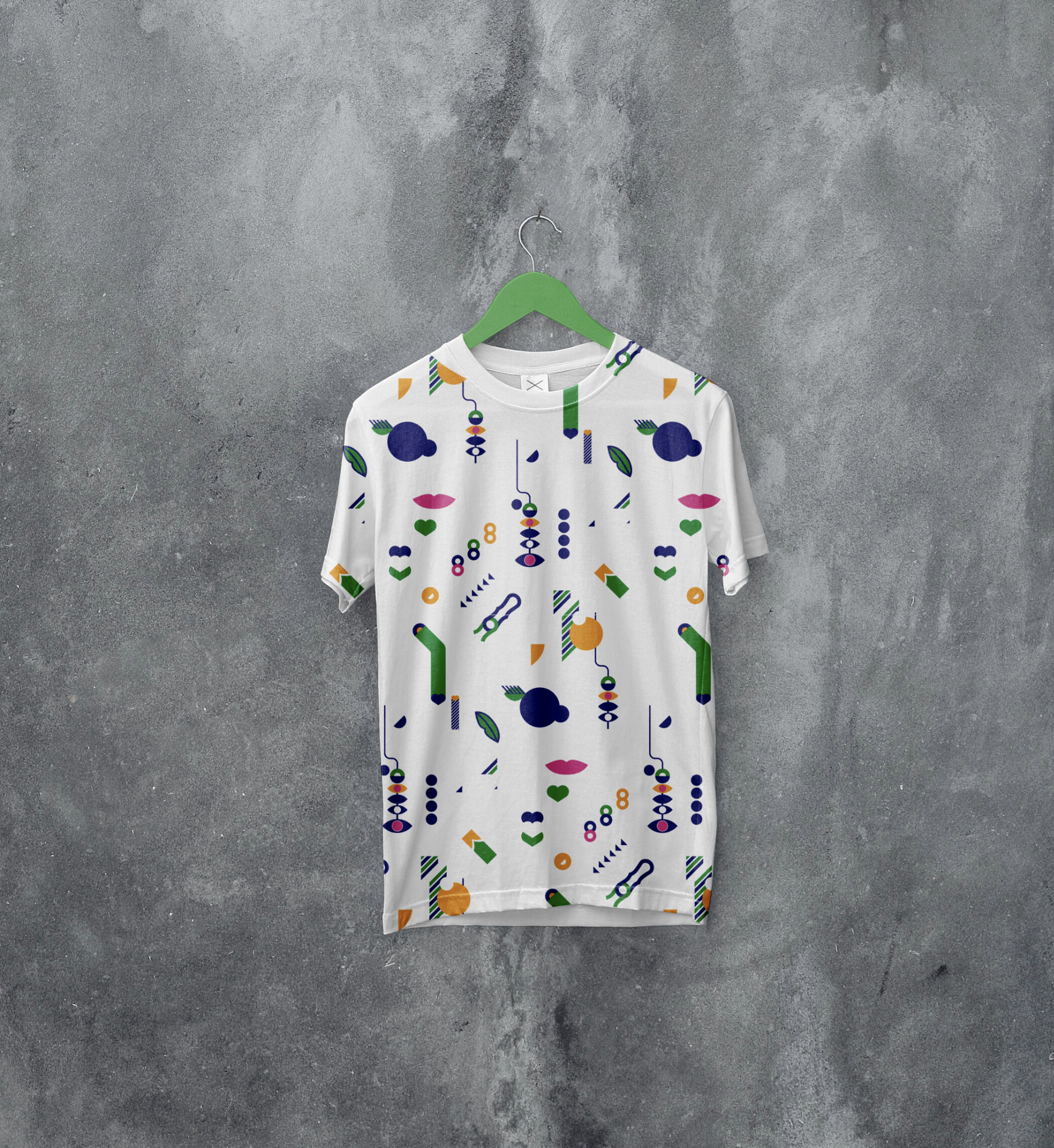 Biała koszulka z krótkim rękawem, wisząca na zielonym wieszaku na tle szarej, betonowej ściany. Na koszulce nadrukowany jest wzór z abstrakcyjnych, kolorowych kształtów.
