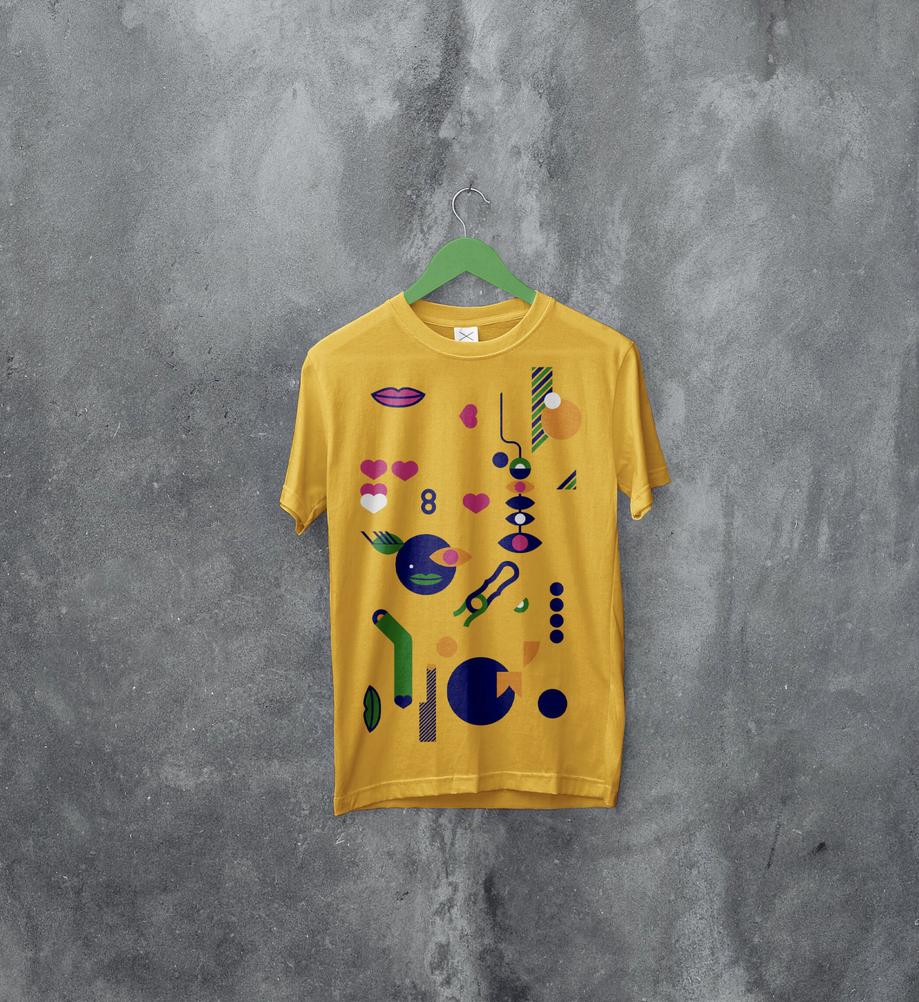 Żółta koszulka z abstrakcyjnym, geometrycznym nadrukiem w kolorach niebieskim, zielonym, pomarańczowym i różowym, zawieszona na zielonym wieszaku na tle szarej, teksturowanej ściany.
