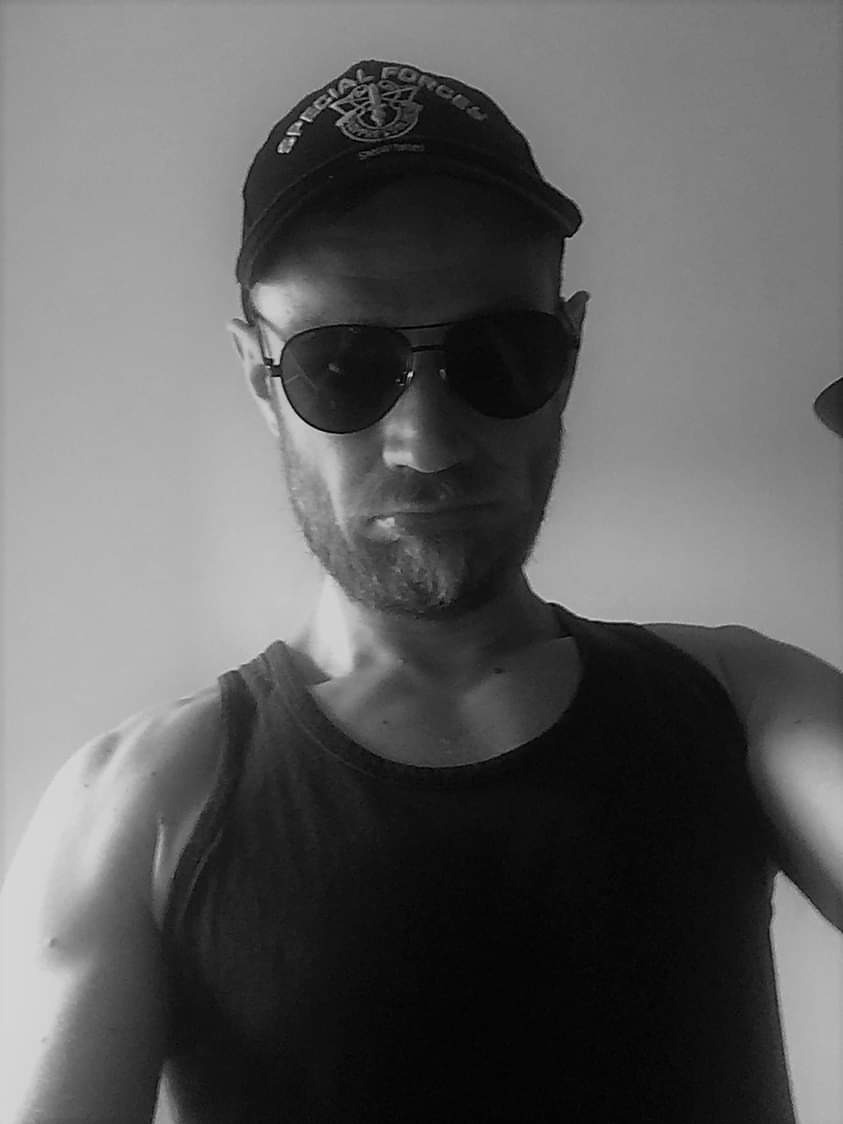 Mężczyzna w czarnej koszulce na ramiączkach i okularach przeciwsłonecznych, z czapką z napisem 'SPECIAL FORCES', portret w czerni i bieli.
