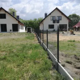 Nowoczesne ogrodzenie panelowe z podmurówką betonową, widoczne na tle dwóch białych domów z ciemnymi dachami, z ziemią wokół w trakcie prac wykończeniowych.