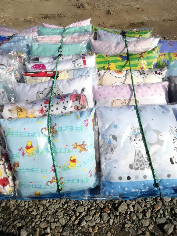 Stos zapakowanych poduszek i poszewek dziecięcych w folii, z różnymi wzorami zwierząt i postaci z bajek, spiętych sznurkiem na kamienistym podłożu.