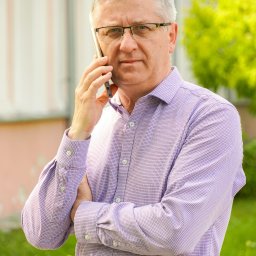 Północ Biuro Nieruchomości Radzyń Podlaski - Mężczyzna w okularach i fioletowej koszuli rozmawia przez telefon, stojąc na zewnątrz na tle budynku i zieleni.