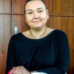 Północ Biuro Nieruchomości Radzyń Podlaski - Portret uśmiechniętej kobiety z włosami upiętymi w kok z różowymi pasemkami, siedzącej za szklanym stołem z kubkiem z napisem 'Dobrze, że jesteś', na tle drewnianej ściany.