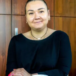 Portret uśmiechniętej kobiety z włosami upiętymi w kok z różowymi pasemkami, siedzącej za szklanym stołem z kubkiem z napisem 'Dobrze, że jesteś', na tle drewnianej ściany.