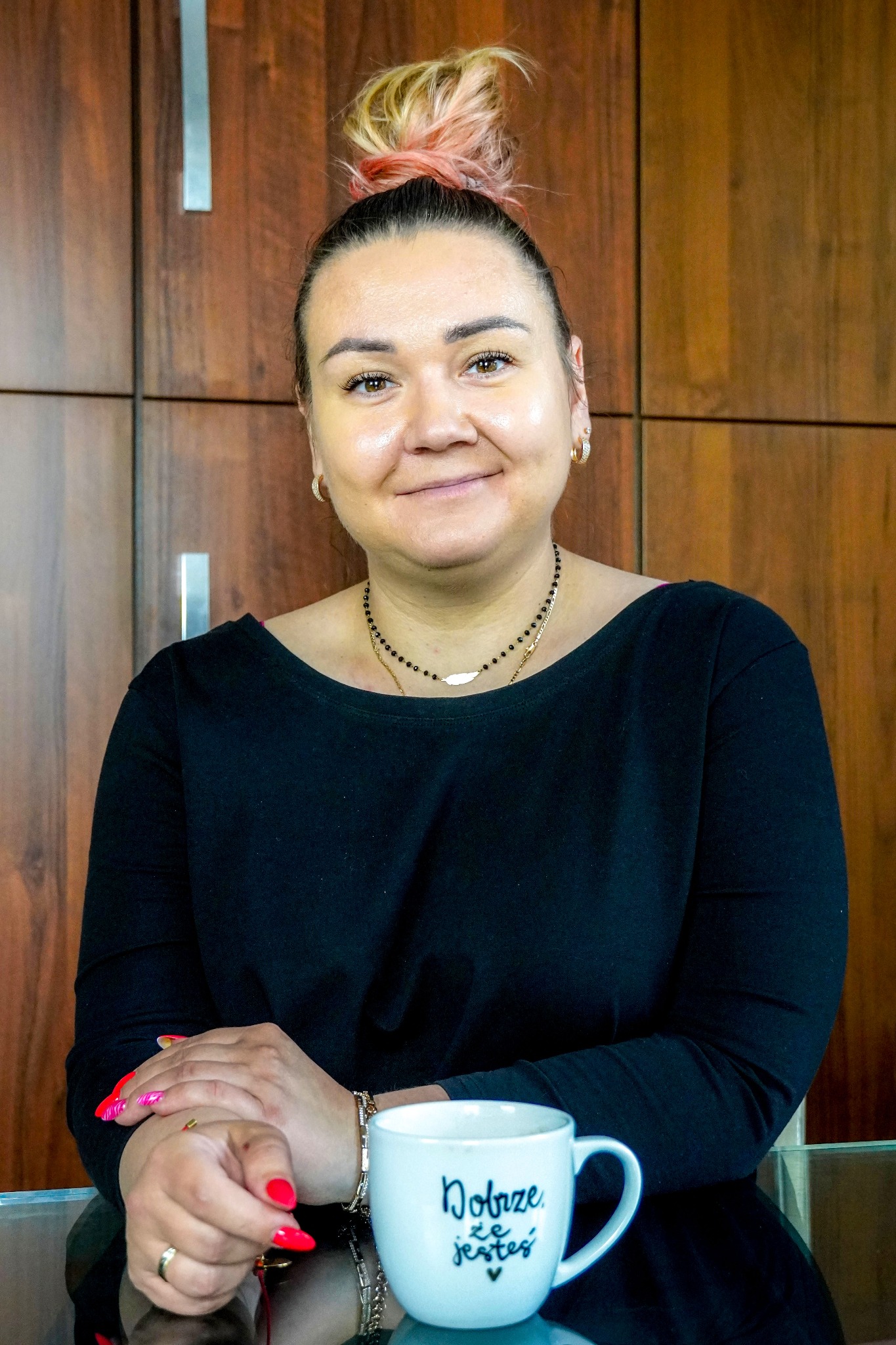 Portret uśmiechniętej kobiety z włosami upiętymi w kok z różowymi pasemkami, siedzącej za szklanym stołem z kubkiem z napisem 'Dobrze, że jesteś', na tle drewnianej ściany.