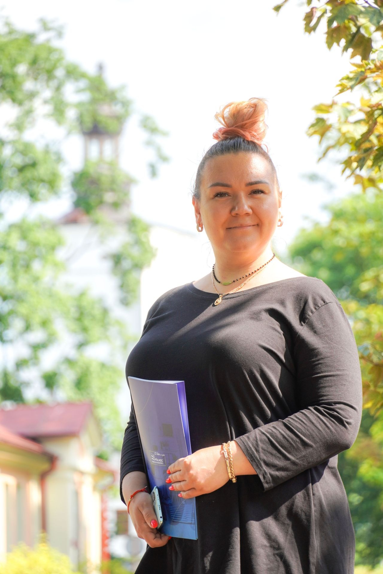 Uśmiechnięta kobieta z upiętymi włosami, ubrana na czarno, trzymająca w dłoniach granatowy folder z grafiką budynku i telefon, na tle zieleni i budynku z czerwonym dachem.