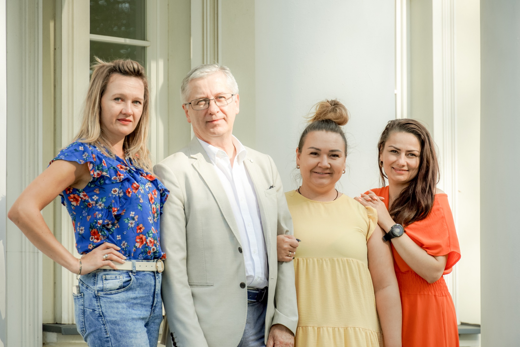 Grupa czterech osób, dwie kobiety po bokach i mężczyzna z dwoma kobietami w środku, pozuje na tle białej kolumnady, sugerując spotkanie zespołu lub konsultacje w biurze nieruchomości.