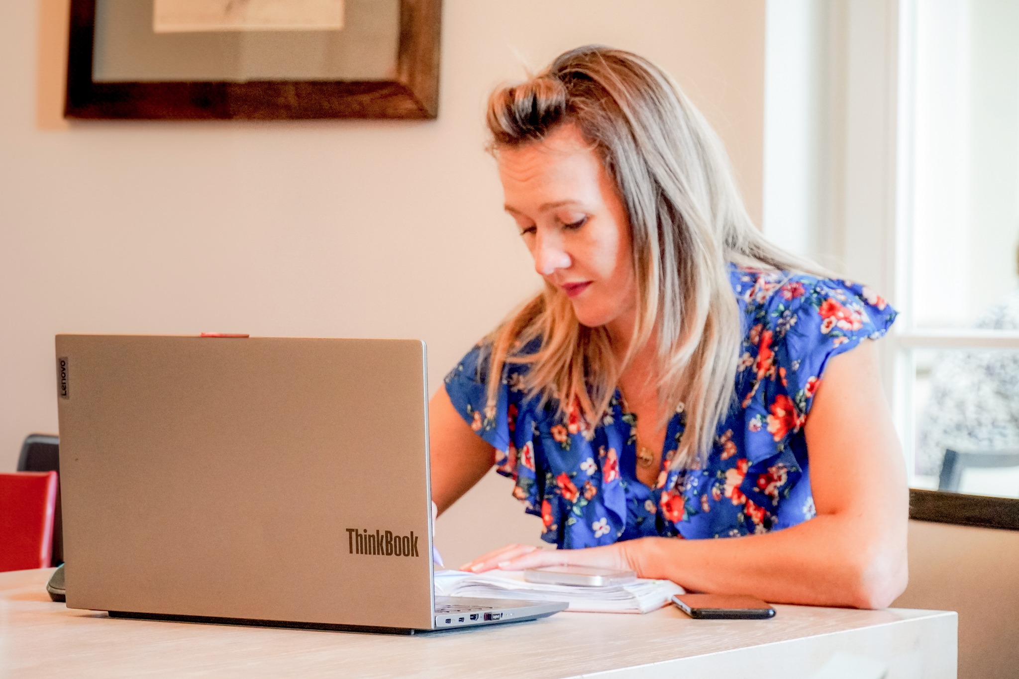 Kobieta w bluzce w kwiaty pracuje przy biurku z laptopem Lenovo ThinkBook, notatkami i telefonem, w jasnym wnętrzu z obrazem na ścianie.