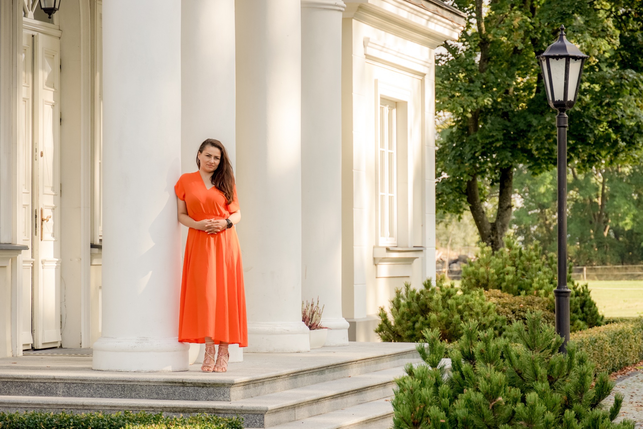Kobieta w pomarańczowej sukience stoi przed białym budynkiem z kolumnami i schodami, w tle zieleń drzew i krzewów, oświetlenie słoneczne.