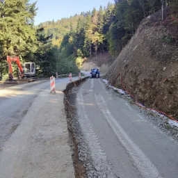 Szeroki kadr na fragment drogi w trakcie budowy, z widocznymi śladami opon, świeżo położonym tłuczniem i pracującym w tle traktorem oraz koparką.