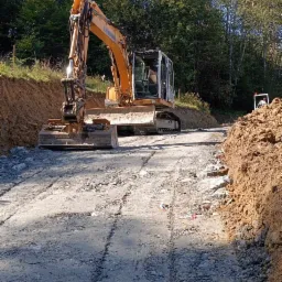 Żółta koparka Liebherr na utwardzonej drodze gruntowej, z widocznymi śladami opon i świeżo wykopanym brzegiem, otoczona zielenią drzew.