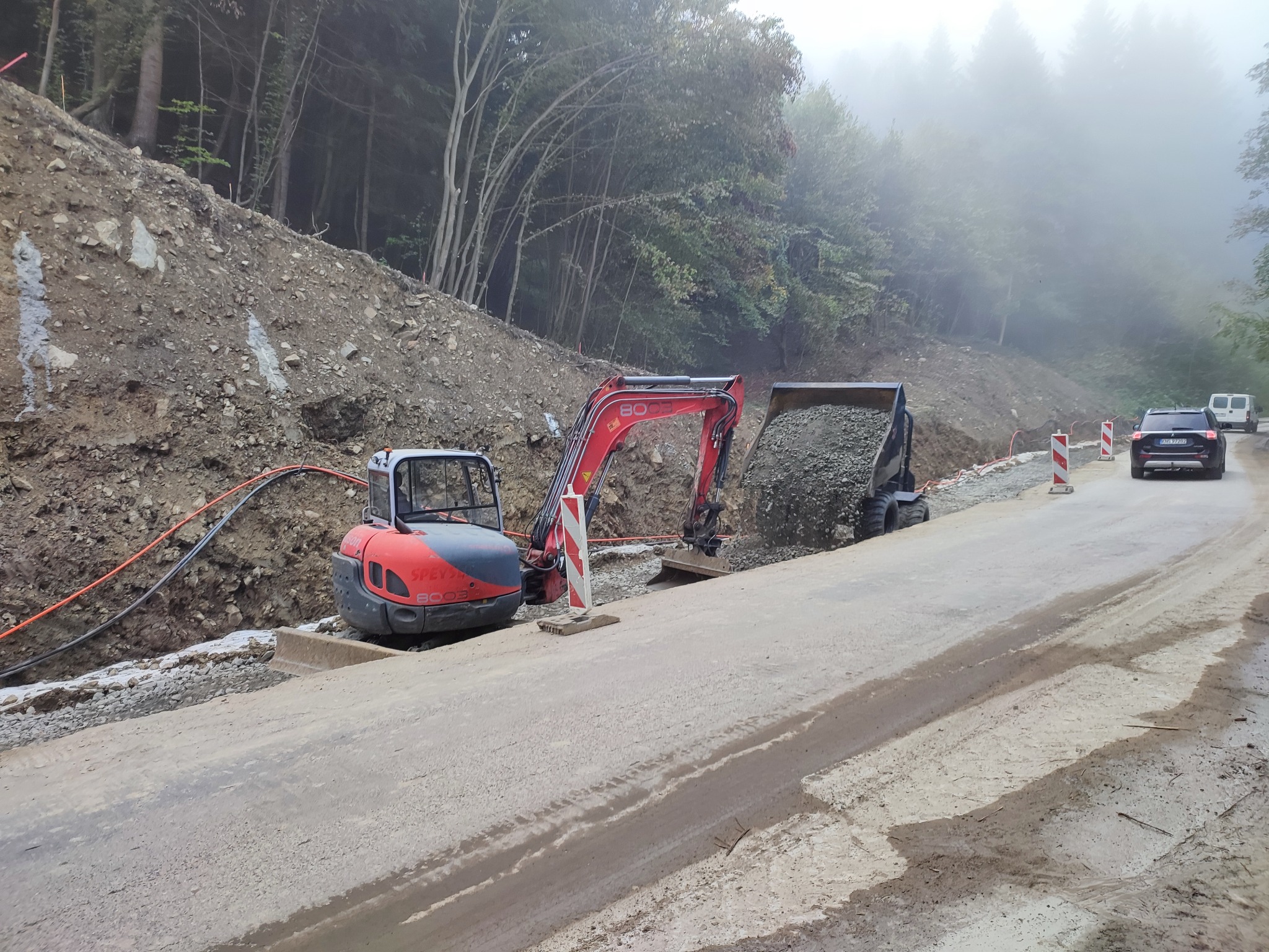 Czerwono-szara mini koparka SPEYSY 8008 ładuje kamienie do wywrotki na tle ziemnego zbocza i zalesionych wzgórz w mglisty dzień, widoczne kable i samochody w oddali.