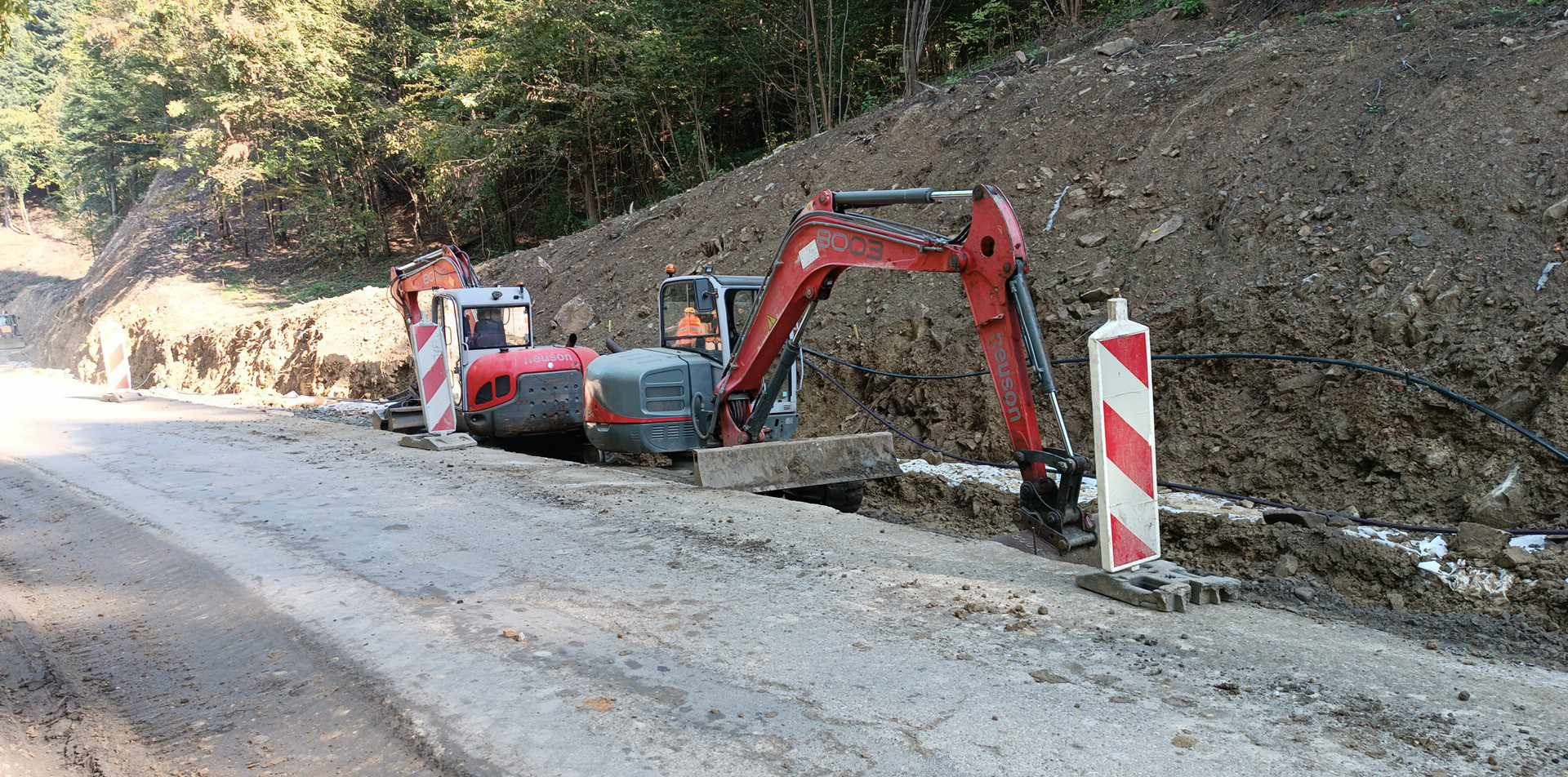 Dwie czerwone mini koparki na betonowej drodze w trakcie prac ziemnych, widoczne przewody w wykopie obok bariery ostrzegawczej.