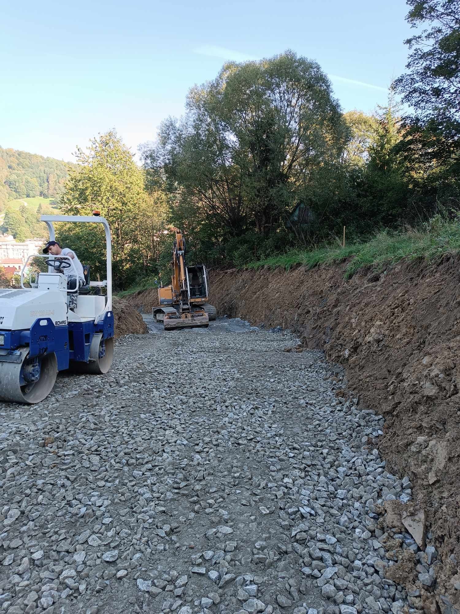 Operator w białej koszulce i czapce obsługuje walec drogowy BOMAG BW 100 AD-4 na żwirowym podłożu, w tle koparka na wykopie, zielone drzewa i zabudowania.