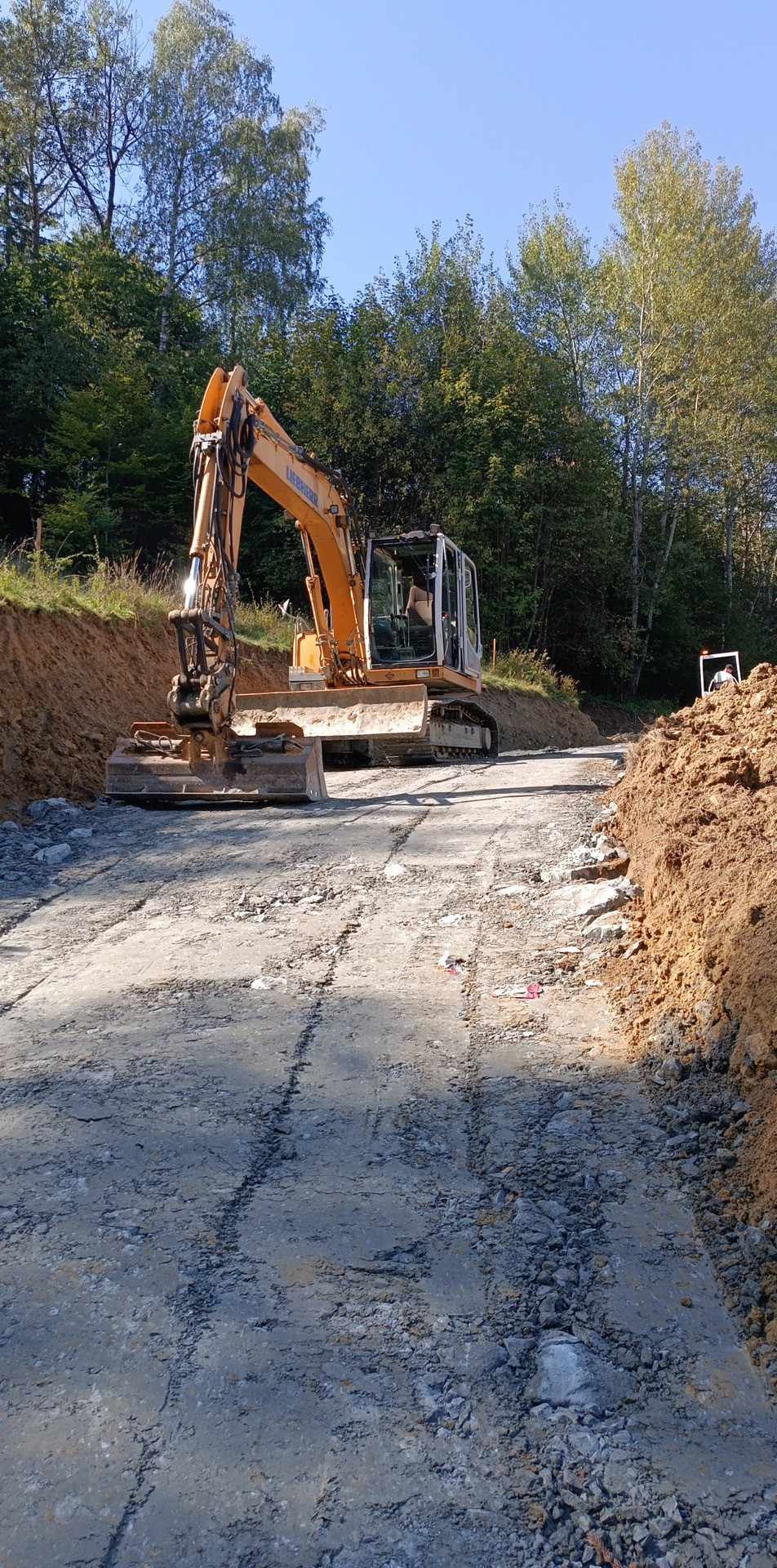 Żółta koparka Liebherr na utwardzonej drodze gruntowej, z widocznymi śladami opon i świeżo wykopanym brzegiem, otoczona zielenią drzew.