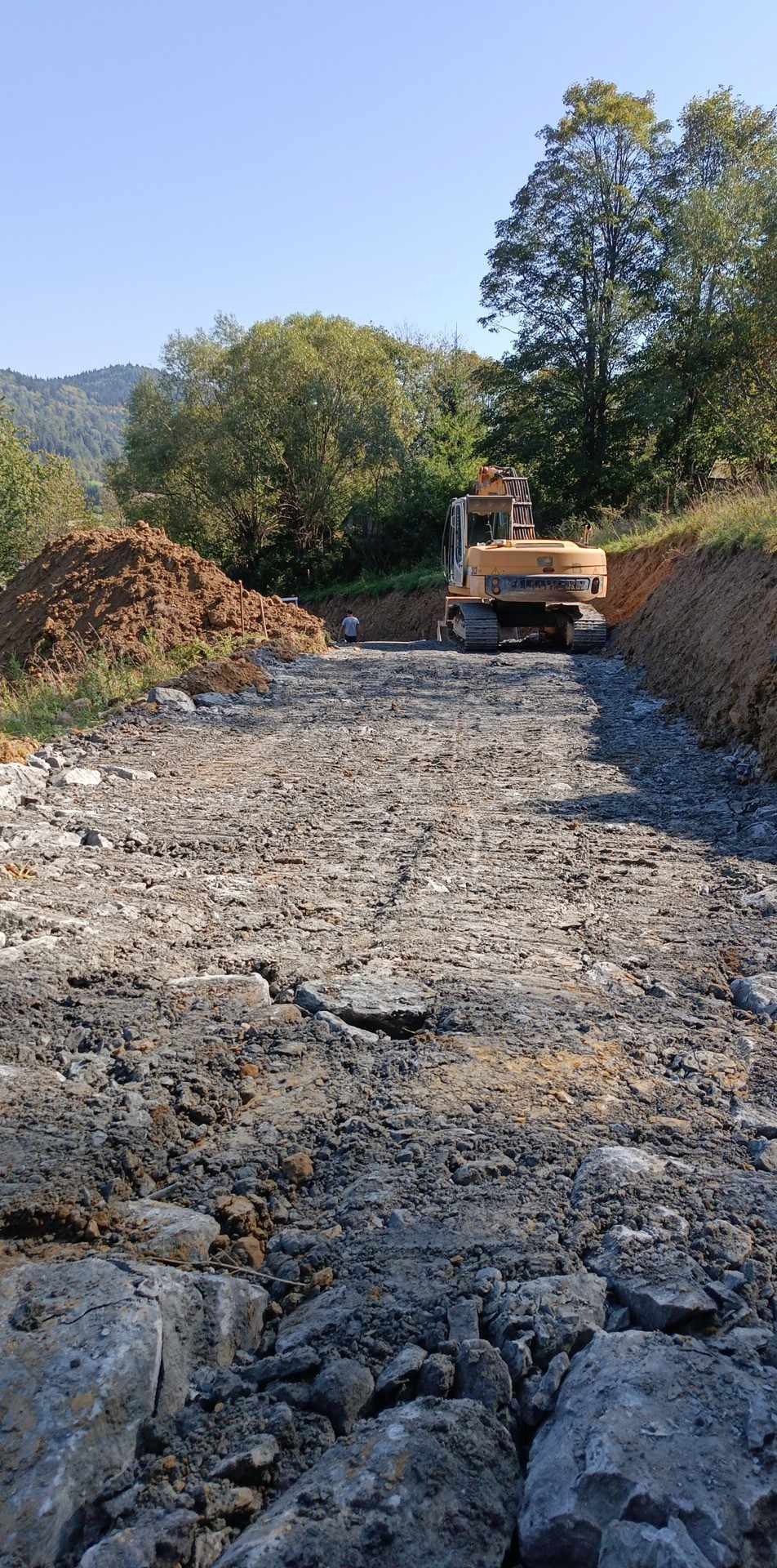 Żółta koparka gąsienicowa na placu budowy drogi, w tle wzgórza i drzewa, widoczny robotnik w oddali.