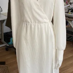 Biała lniana sukienka z długim rękawem i kopertowym dekoltem na manekinie krawieckim w pracowni, widoczna maszyna do szycia i półki z akcesoriami w tle.