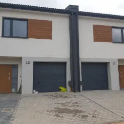 Nowoczesne domy bliźniacze z jasną elewacją, drewnianymi akcentami i szarymi bramami garażowymi, numerami 10 i 12, oraz kostką brukową na podjeździe.