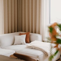 Szyra Studio - Jasny narożnik z poduszkami w odcieniach beżu i brązu, przykryty kremowym pledem o wyraźnej fakturze, na tle okna z beżowymi zasłonami.