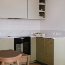 Szyra Studio - Nowoczesna kuchnia z oliwkowymi szafkami, marmurowym blatem i drewnianą podłogą; na blacie misa z owocami i ceramiczne gniazdka elektryczne.