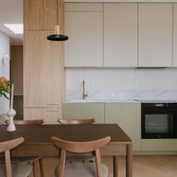 Nowoczesna kuchnia z drewnianym stołem i krzesłami, kwiaty w ozdobnym wazonie na blacie, minimalistyczne szafki w odcieniach beżu i zieleni, złota armatura i lampa wisząca.