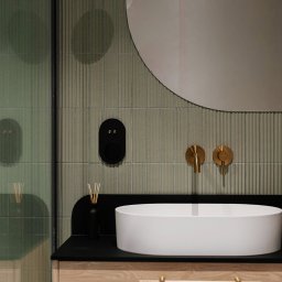 Szyra Studio - Nowoczesna łazienka z owalną umywalką nablatową, złotą armaturą, zielonymi płytkami ściennymi z subtelnym wzorem i okrągłym lustrem bez ramy.
