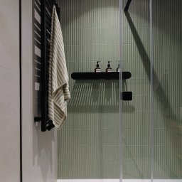 Szyra Studio - Nowoczesna łazienka z kabiną prysznicową ze szklanymi drzwiami, zielonymi, pionowo żłobionymi płytkami, czarnym grzejnikiem drabinkowym z zawieszonym ręcznikiem w paski oraz półką z trzema...